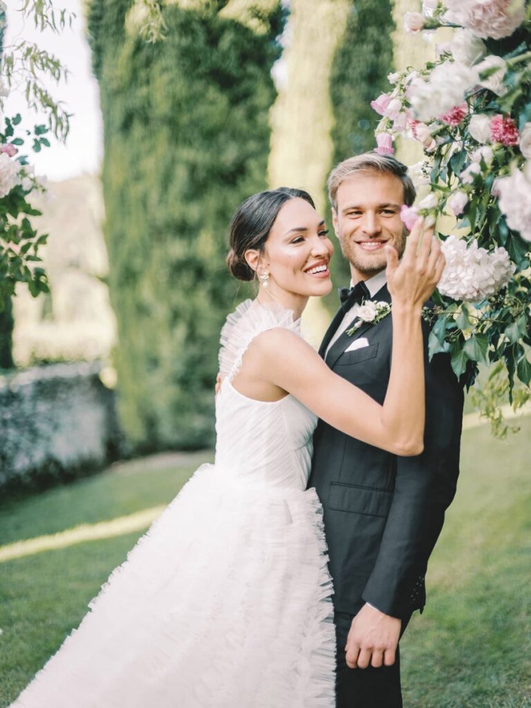 A joyful couple in wedding attire stands in villa Cetinale. The bride wears a ruffled white gown, and the groom is in a black suit, surrounded by pink florals.