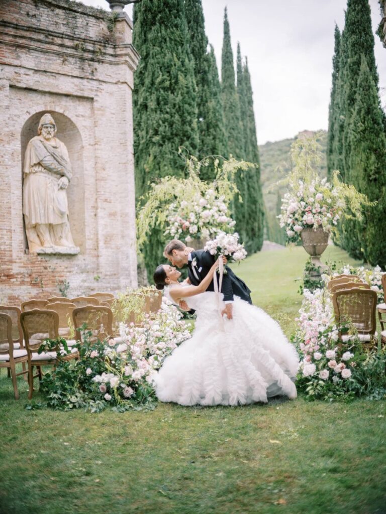 A couple embraces in a romantic dance in villa Cetinale for their garden wedding, surrounded by lush pink and white flowers. Tall cypress trees and an ancient statue form the backdrop.