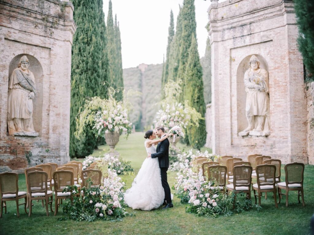 A bride and groom embrace in front of the ceremony setup in villa Cetinale, surrounded by chairs and floral arrangements. Tall stone statues and cypress trees frame the scene.