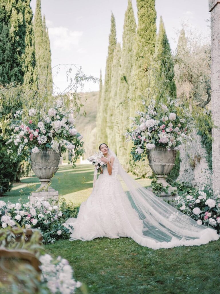 A bride in a flowing white gown and veil stands amidst lush greenery in villa Cetinale.