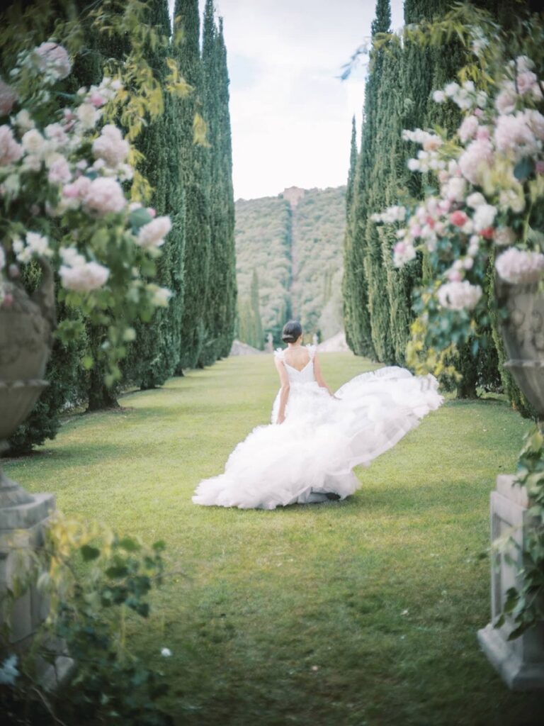 A bride in a flowing white gown and veil stands amidst lush greenery in villa Cetinale.