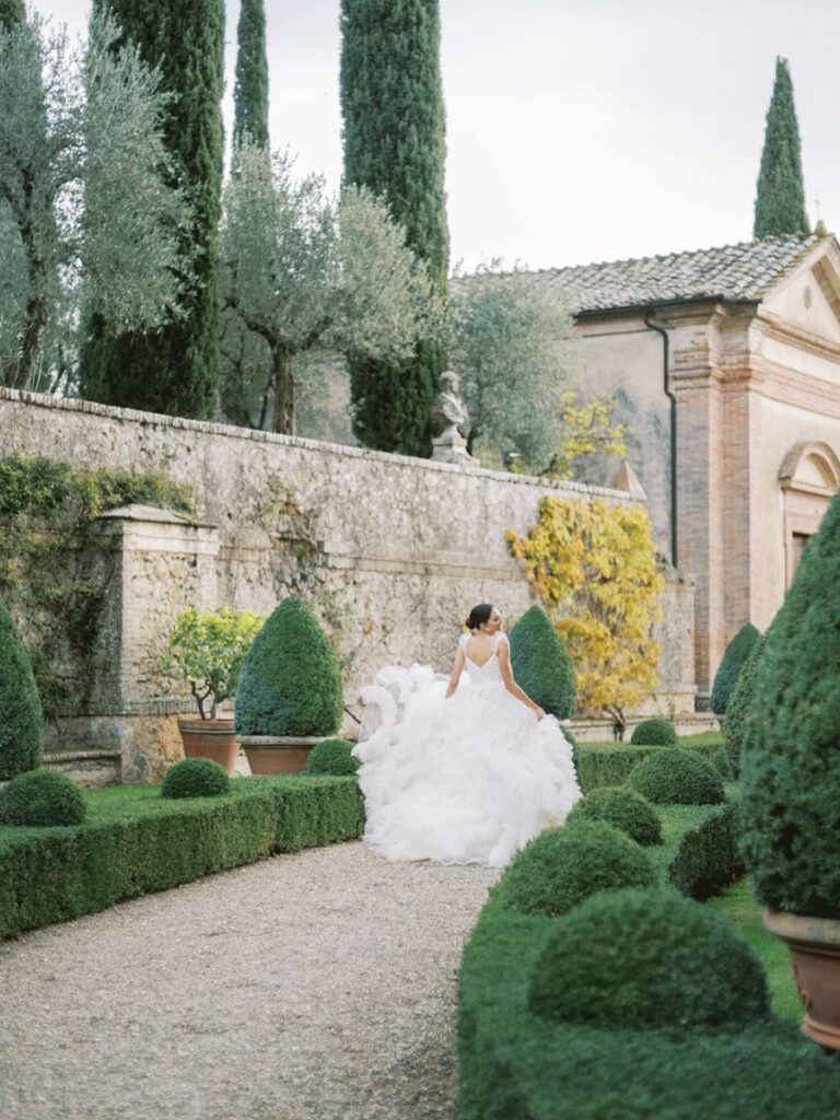 Bride in voluminous white dress walks through a lush garden path in villa Cetinale in front of the chapel.