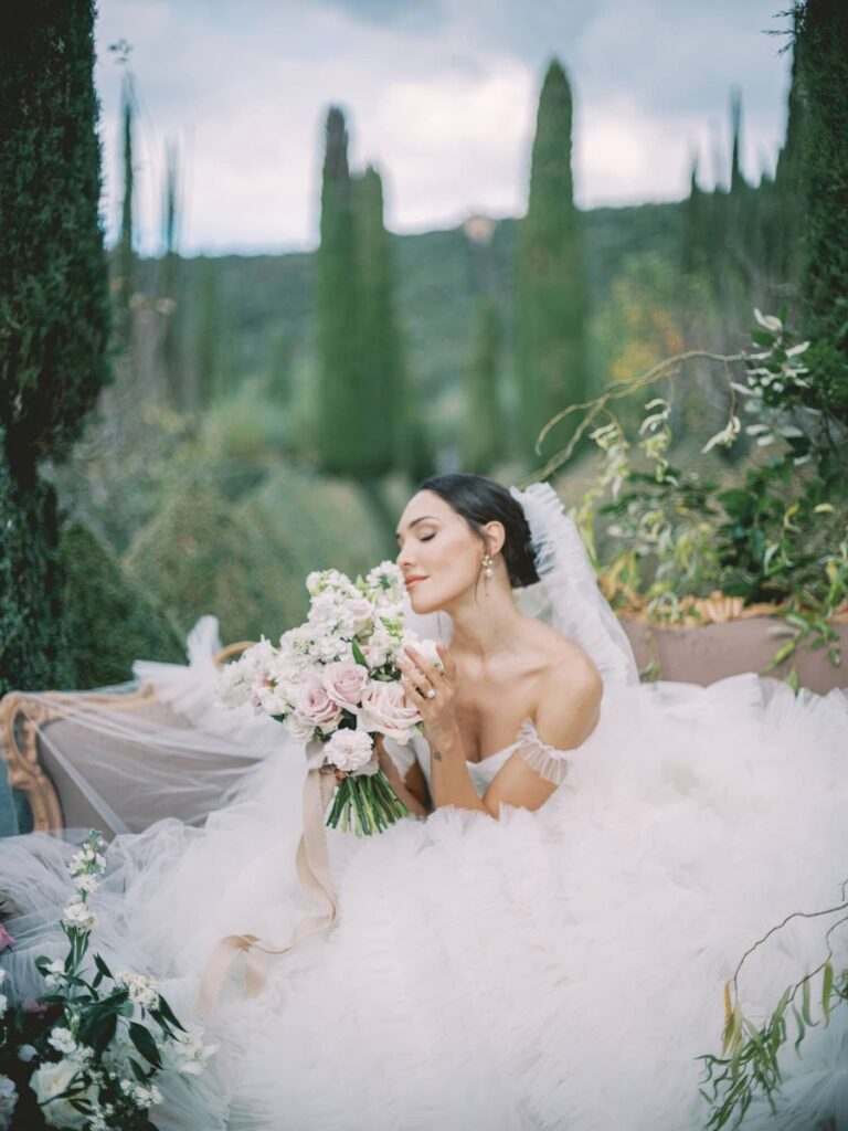 Bride in villa Cetinale in a voluminous white gown gently holds a bouquet of pink and white flowers, surrounded by lush greenery, conveying elegance and serenity.