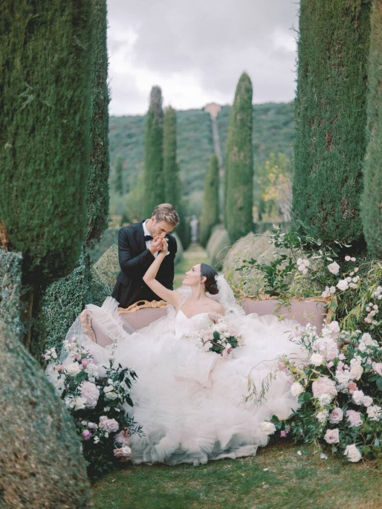 A couple in villa Cetinale surrounded by pink and white flowers. A groom in a suit gently kisses her hand, exuding romance amidst towering green trees.