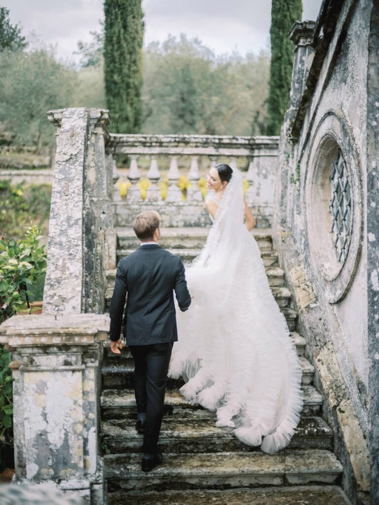 A bride in a white, flowing gown ascends an old stone staircase in villa Cetinale, holding hands with a groom in a black suit. The scene is serene and romantic.