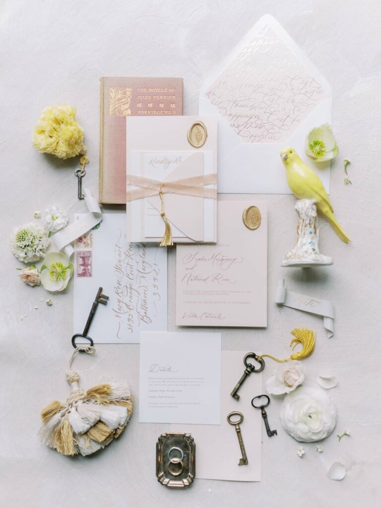 A vintage villa Cetinale wedding flat lay with elegant invitations, a pink book, brass keys, a yellow bird figurine, and soft flowers on a textured white background.