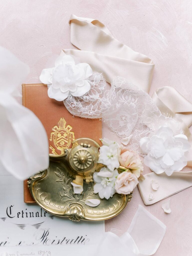 Elegant flat lay with a vintage brass tray, soft peach roses, white flowers, lace, and a beige ribbon on a pink surface, conveying a romantic tone.