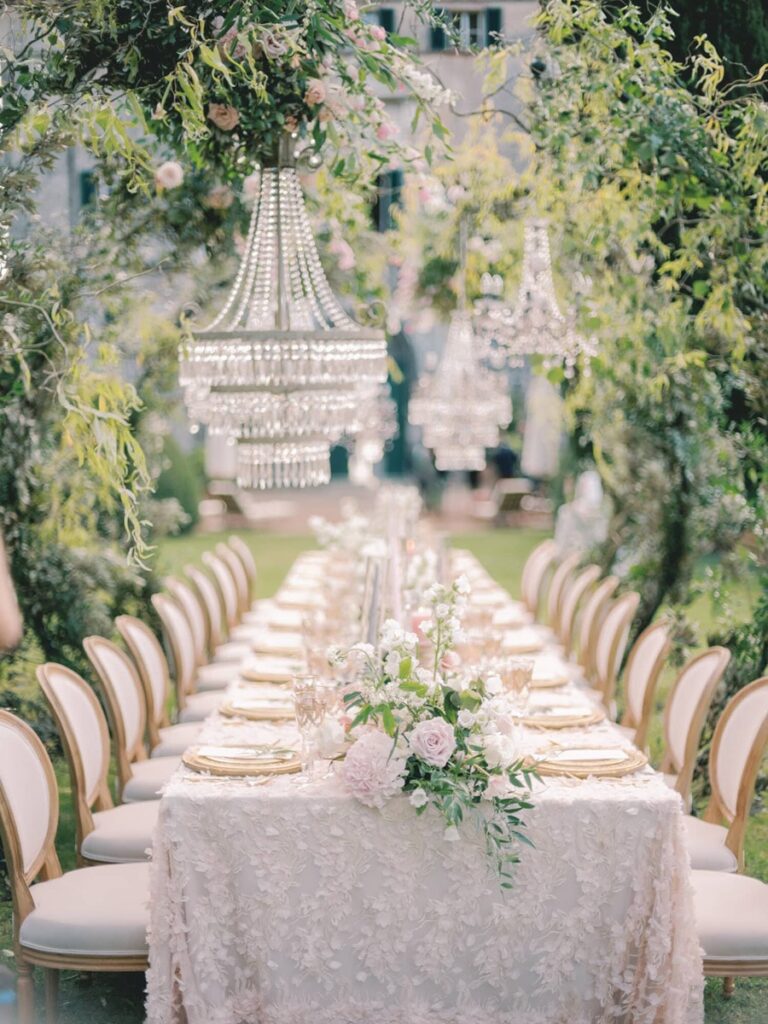 Elegant outdoor dining scene in villa Cetinale with a long table adorned with floral lace, pastel flowers, and crystal chandeliers hanging from lush greenery, creating a romantic ambiance.