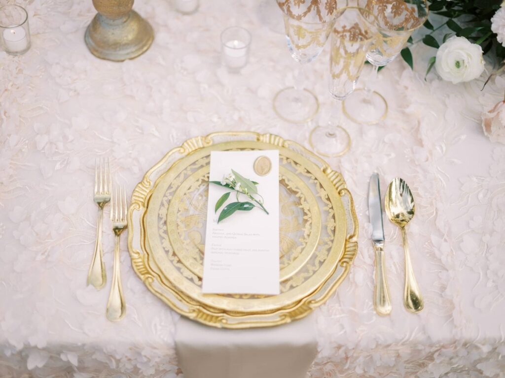 Elegant table setting with a gold-rimmed plate, floral menu card, and gold cutlery on a textured white tablecloth. A refined, festive ambiance.