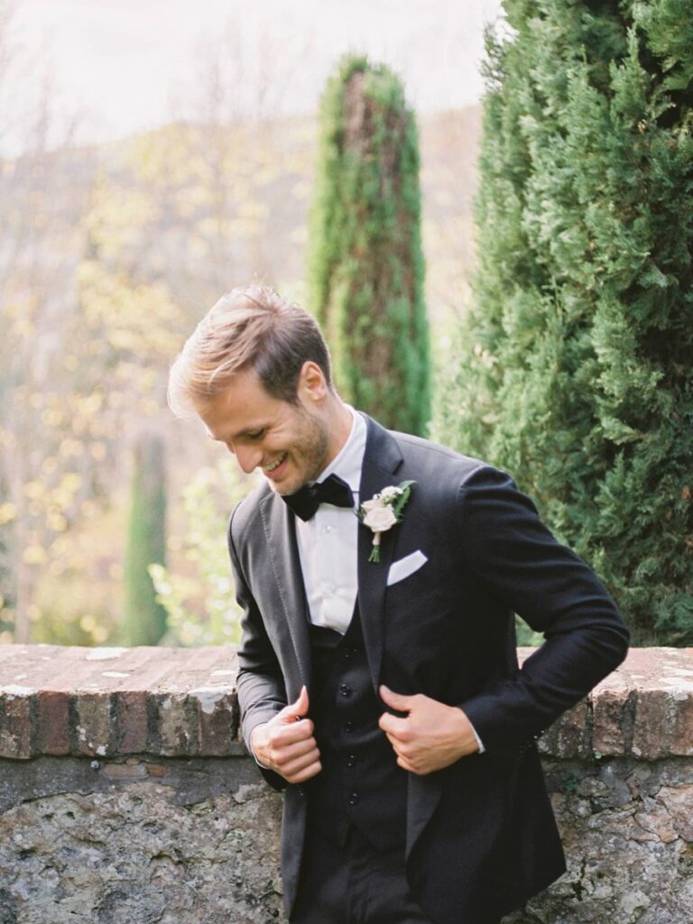 Smiling Cetinale groom in a tuxedo leans against a rustic stone wall, with lush greenery and blurred trees in the background, conveying a joyful, serene mood.