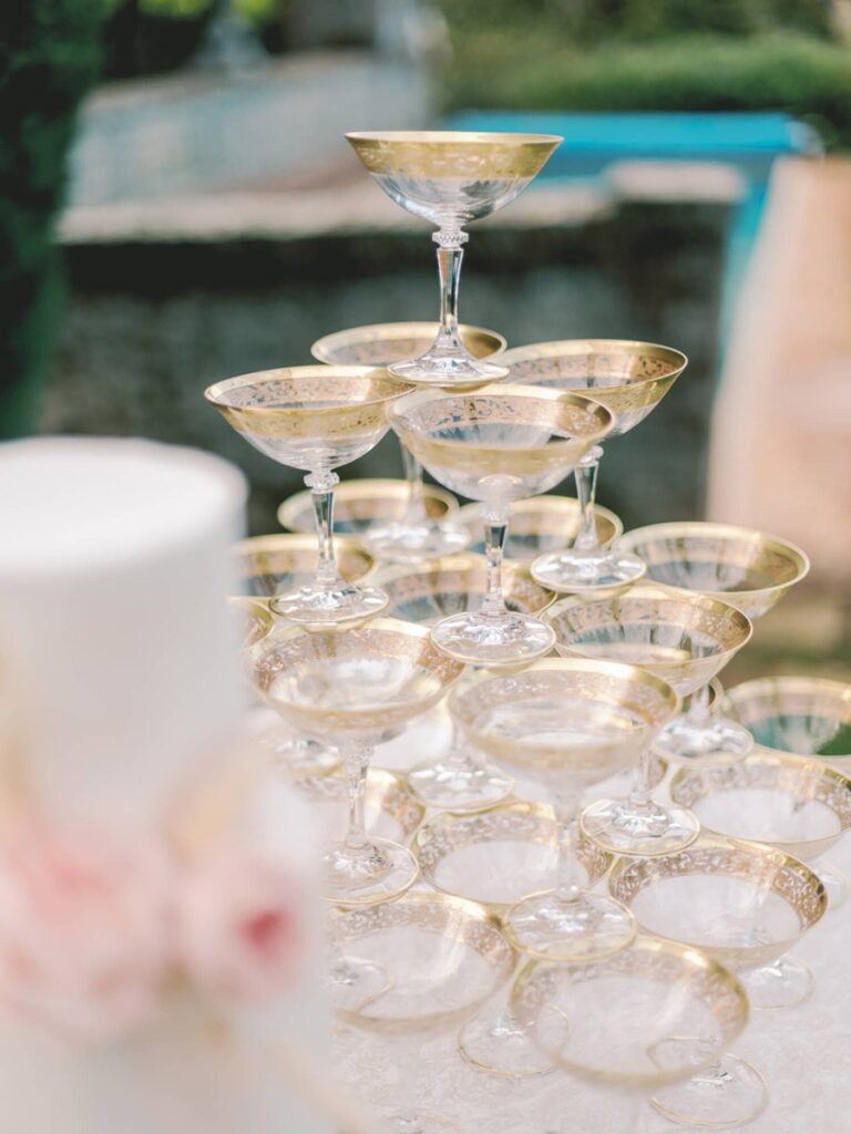 A pyramid of elegant champagne glasses with gold rims is set up outdoors, conveying a festive and luxurious atmosphere. A blurred white and pink element is in the foreground.