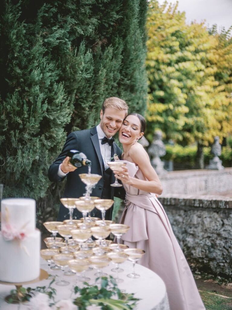 A joyful couple at a wedding pouring champagne into a tower of glasses in villa Cetinale. They're dressed elegantly, surrounded by lush greenery and a wedding cake.