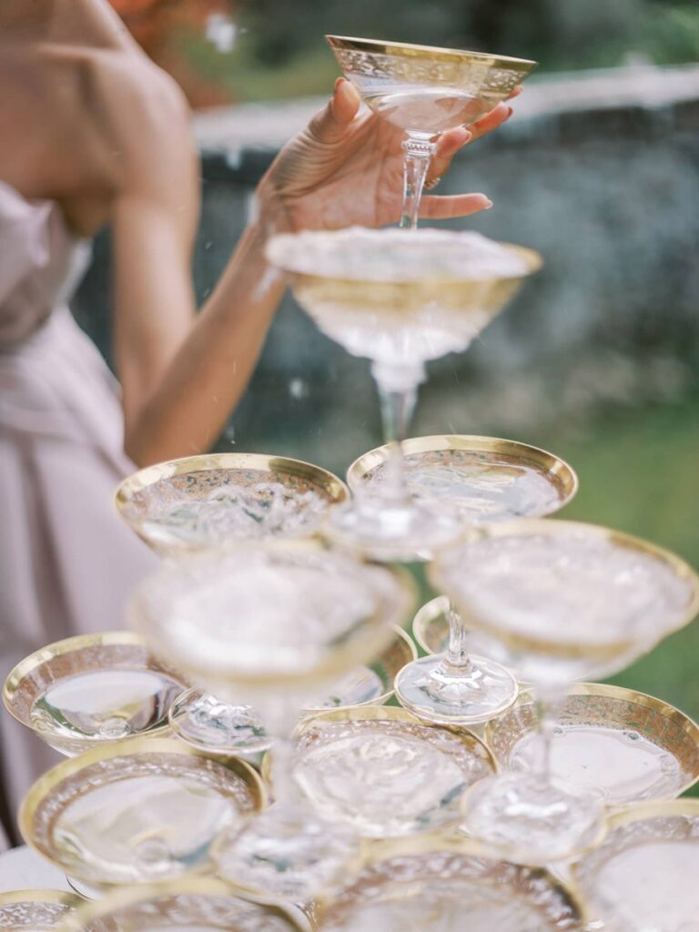 Bride's hand holds a champagne glass atop a cascading tower of glasses, each filled with bubbly liquid. Elegant and celebratory atmosphere outdoors.