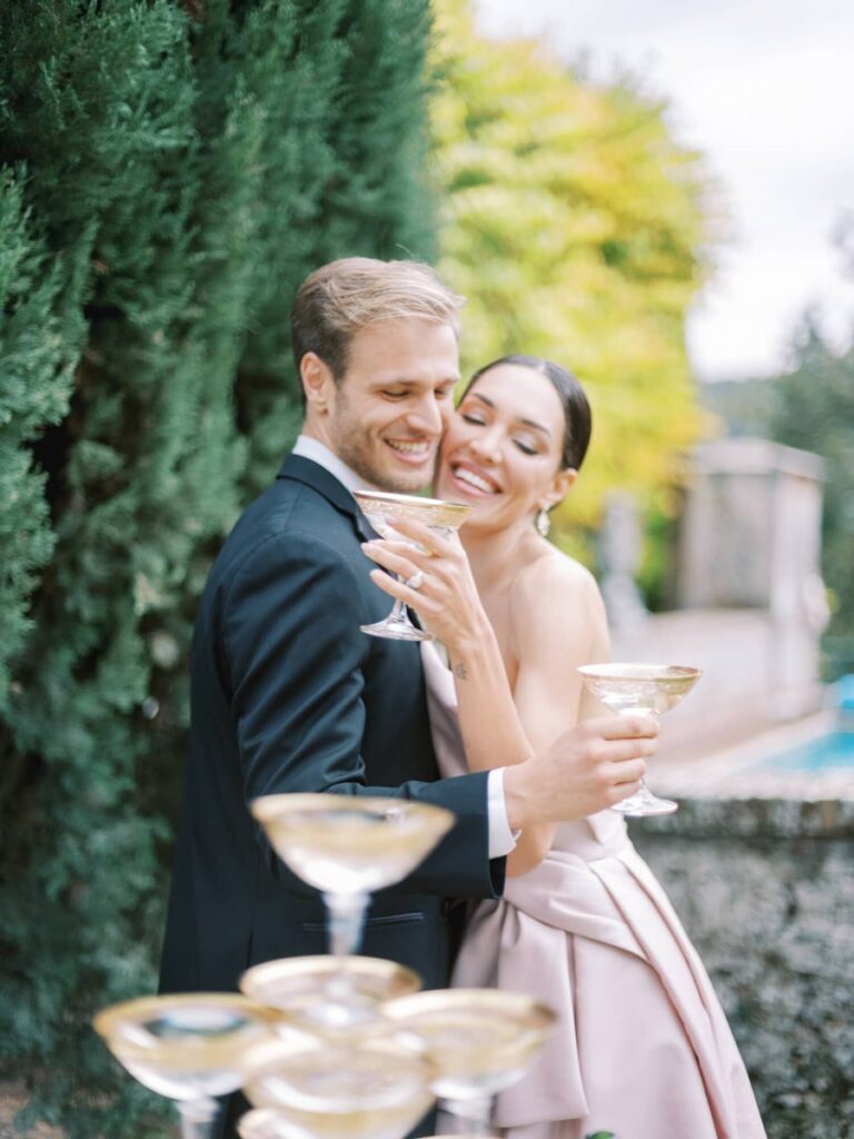 A couple kisses while the man holds a champagne glass. He is in a black tuxedo, and she is in a strapless gown. The setting is outdoors, surrounded by tall greenery, conveying romance.