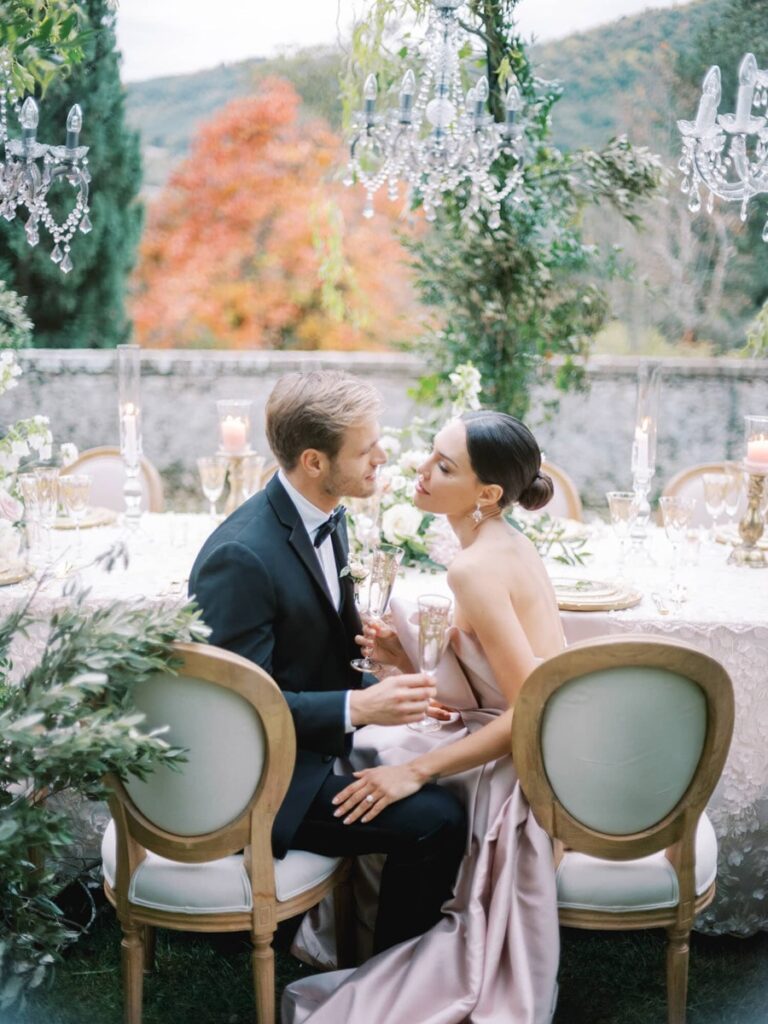 A couple sits intimately at a lavishly decorated outdoor table with chandeliers, surrounded by greenery and autumn foliage, exuding romance and elegance.