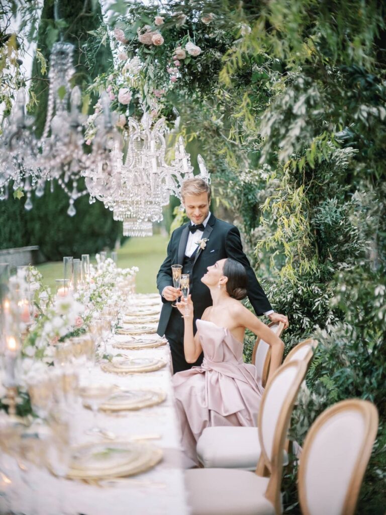 A couple elegantly toasts at an outdoor table adorned with flowers and candles. Crystal chandeliers hang above, creating a romantic, sophisticated atmosphere.