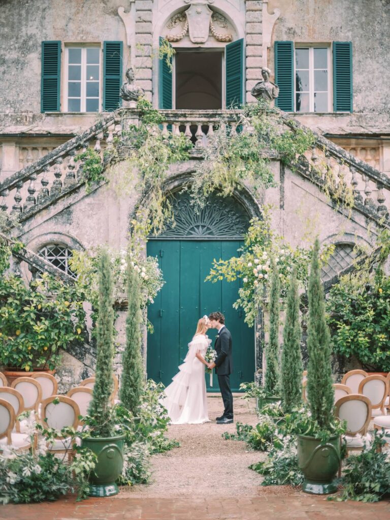 A couple kisses in a romantic garden wedding setting in front of villa Cetinale, with vintage chairs, green foliage, and a rustic building backdrop, conveying an elegant, serene tone.