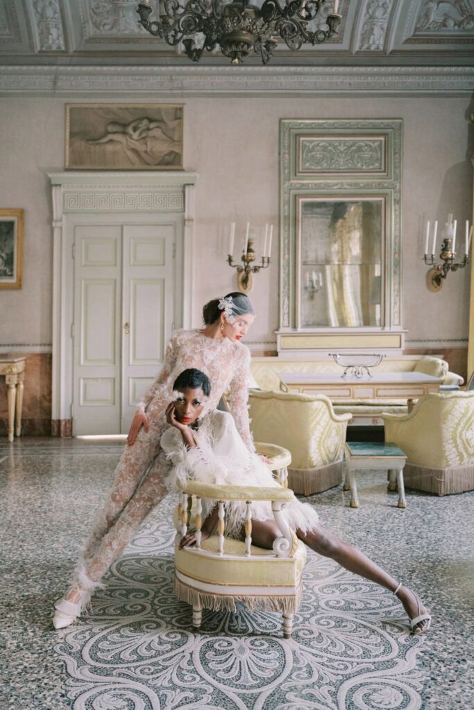 Two elegantly dressed models pose in an opulent room in villa Pizzo, featuring intricate patterns and soft lighting.